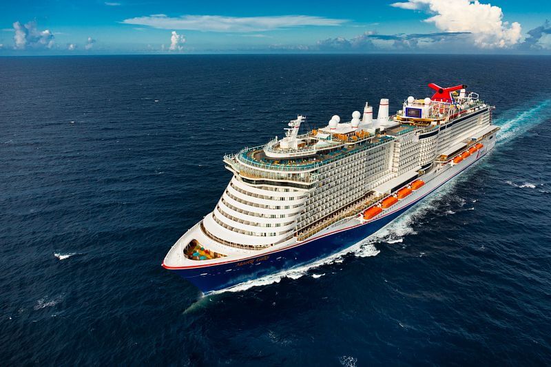 The image shows the Carnival Mardi Gras cruise ship sailing through open ocean, leaving a light trail of wake behind it. The ship’s distinct red and blue hull stands out, with multiple decks featuring balconies, pools, and recreational areas. The clear blue sky and expansive sea emphasize the grandeur and luxury of the vessel on its journey.