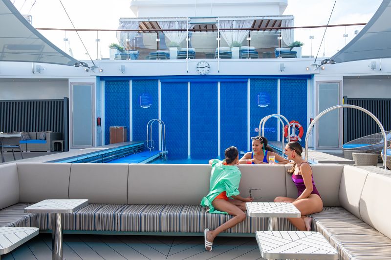 A group of cruise guests is relaxing at the Serenity area on Carnival's Mardi Gras. Three women in swimsuits are seated on a striped couch and at a nearby table, engaged in conversation and enjoying drinks. In the background, a serene pool area with blue tiles and deck chairs is visible, along with an upper deck featuring cabanas and potted plants, creating a tranquil and luxurious ambiance.
