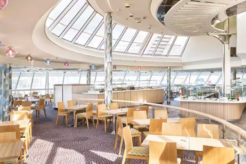 The image depicts the Marella Cruises Islands Buffet. The dining area features light wood tables and chairs, with large windows and skylights allowing ample natural light. The modern design includes geometric-patterned columns and a clean, open layout with various food stations in the background.