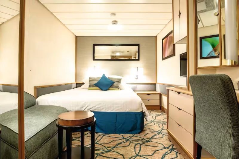A cozy single cabin on a Marella cruise ship featuring a neatly made bed with white linens and blue accent pillows. The room includes a small sitting area with a round table, a dresser with a mirror, and a TV mounted on the wall, all accented with warm, neutral tones and modern decor.