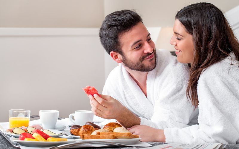 A couple in white bathrobes enjoys room service on Marella Cruises. The breakfast tray includes croissants, pastries, fruit, and beverages like coffee and orange juice. They are smiling at each other, creating a cozy and intimate atmosphere.