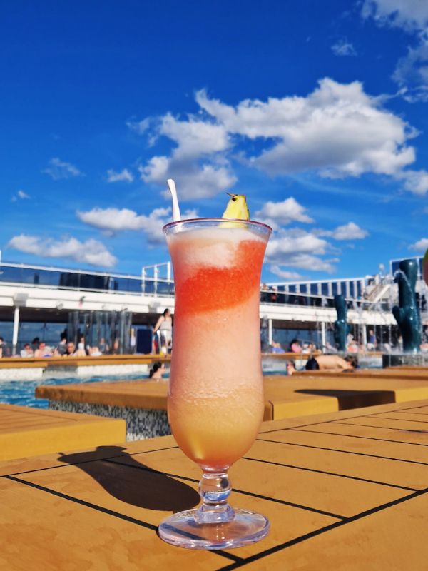 A Miami Vice cocktail on a Carnival Cruise ship, featuring vibrant layers of red and yellow slush with a pineapple garnish. The drink is set on a wooden poolside table with passengers lounging and swimming in the background under a bright blue sky dotted with fluffy white clouds.