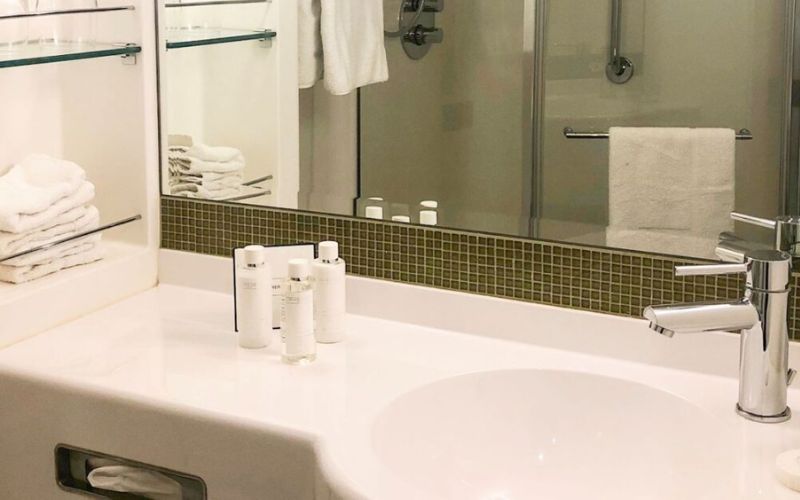 Spacious cruise ship bathroom featuring a clean white sink with miniature toiletry bottles lined up on the counter, a glass shower stall in the background, and neatly folded white towels stacked on the shelves.