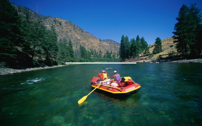 The image captures an exhilarating moment of two people river rafting in a bright red raft, navigating through a scenic river with lush, mountainous terrain in the background. Both individuals are equipped with life jackets and paddles, actively rowing through the vibrant green waters. This picturesque setting emphasizes the thrilling experience of outdoor adventure sports, perfect for those seeking an adventurous escape in nature.