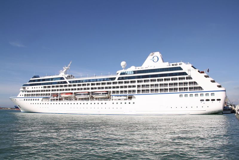Cruise ship Nautica, captured on a bright day with clear blue skies. The ship is presented in a profile view, showcasing its multi-deck structure with rows of balconies and windows, painted predominantly in white. The vessel's name is visible on the hull, and it sports a large satellite dome and other navigational equipment on top. The tranquil sea around it emphasizes the leisurely atmosphere of a cruise setting.