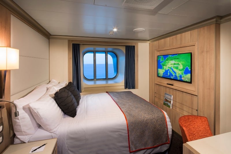 A cozy solo cabin on the HAL Nieuw Statendam cruise ship, featuring a comfortable bed with white linens and dark accent pillows. The room includes a large window with ocean views, a wall-mounted flat-screen TV, and a small desk with an orange chair. The decor combines warm wood tones with modern furnishings, creating a welcoming and elegant atmosphere.