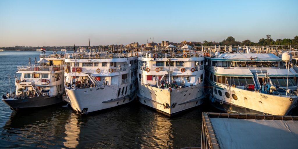 Several Nile cruise ships moored side by side at a dock in the golden hour of the early evening. The ships are multi-decked with rows of windows and life rings, bustling with activity against a backdrop of the distant shoreline under a clear sky. This scene captures the vibrant atmosphere of river cruising in Egypt.