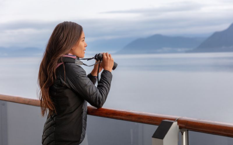 A woman on a cruise ship gazes out at the calm sea and distant mountains through binoculars, dressed warmly for cool weather. The sky is overcast, adding a serene and peaceful atmosphere to the scene.