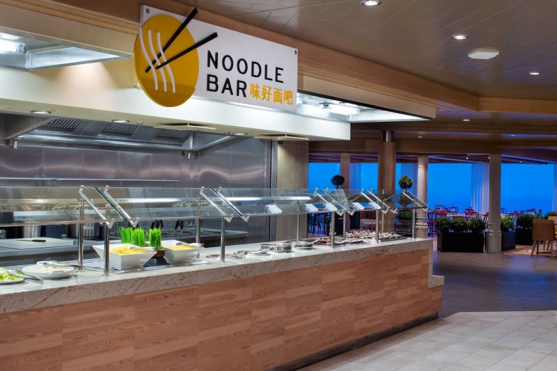 The Noodle Bar on Royal Caribbean's Spectrum of the Seas, featuring a well-lit, clean serving counter with various ingredients on display, and the sign 'NOODLE BAR' in English and Chinese characters overhead, inviting guests to a culinary experience with a view of the ocean through the windows.