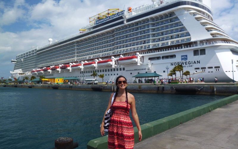 A woman in a red and white summer dress stands smiling at the dockside with the Norwegian Breakaway cruise ship moored in the background, under a bright blue sky with some clouds.