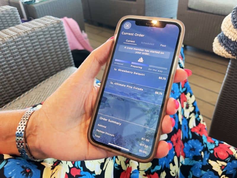 A person using the OceanNow app on a Princess cruise ship to check the status of their current order. The app screen shows that a crew member has started preparing a Strawberry Daiquiri and an Ultimate Pina Colada, with an order summary listing the subtotal and total as $0.00 due to a package discount. The person's hand is holding the phone, and they are seated in an area with wicker furniture.