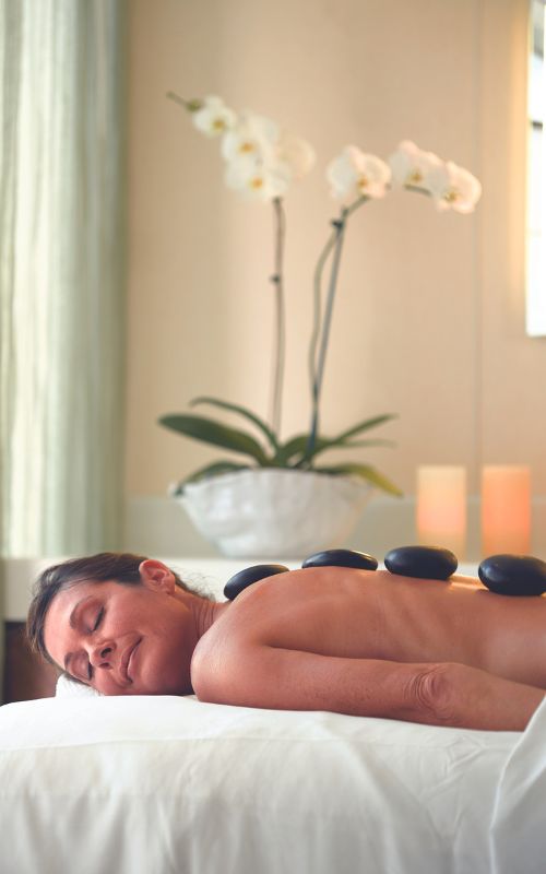 A woman receiving a hot stone massage in a serene spa setting, with white orchids and soft lighting in the background. She appears relaxed and at peace on the massage table.