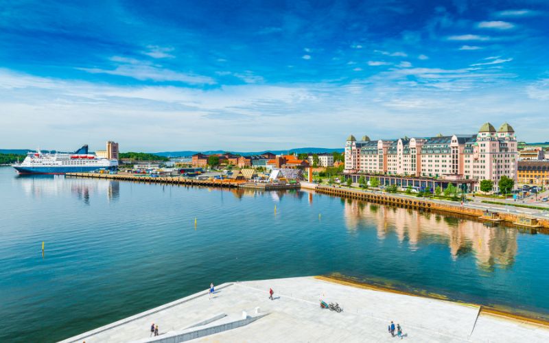The vibrant waterfront of Oslo, Norway, highlighting the harmonious blend of historical architecture and modern development. A cruise ship is moored at the port, adding a dynamic element to the cityscape. The waterfront buildings, with their colorful facades and distinctive European architectural styles, reflect Oslo's rich cultural heritage. The foreground shows a wide, open promenade that appears to be a popular spot for both locals and tourists, providing a leisurely space for walks with a view of the fjord. The backdrop of gentle hills and a clear sky enhances the picturesque setting, showcasing Oslo as a city where historical charm meets contemporary living.