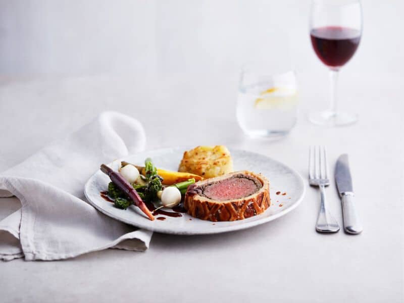 A gourmet plate of Beef Wellington served with roasted vegetables, a potato gratin, and a drizzle of sauce on P&O Main Dining Room. Accompanied by a glass of red wine and a glass of water with a lemon slice in the background, the setting exudes fine dining.
