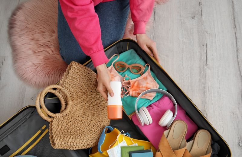 A person in a pink sweatshirt packs a suitcase with summer essentials, including a straw bag, sunscreen, sunglasses, a bikini, headphones, sandals, and travel documents. The suitcase is laid open on a wooden floor with a pink fuzzy rug nearby, suggesting preparation for a sunny vacation.