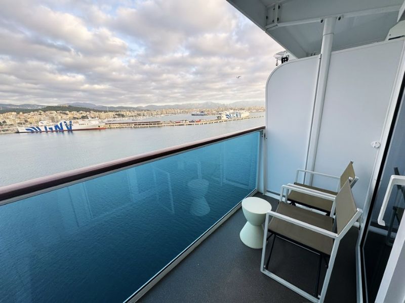 A view from the balcony of a cruise ship docked at the port of Palma de Mallorca. The balcony features a simple set-up with a pair of metal-framed chairs and a small table, perfect for enjoying the scenic views. The railing of the balcony provides an unobstructed view of the bright blue waters of the harbor and the cityscape of Palma in the distance. The overcast sky adds a tranquil, soft light to the scene, making it ideal for relaxation while taking in the beauty of this popular Mediterranean destination.