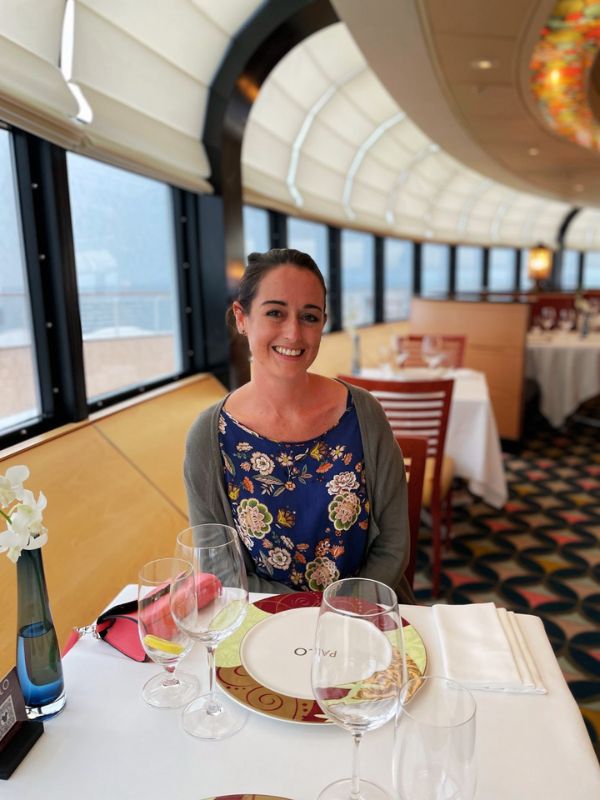 Cruise Mummy seated at a table in the Palo restaurant aboard the Disney Magic cruise ship. The restaurant's interior features large arched windows offering a panoramic ocean view, contributing to a bright and airy atmosphere. The table is elegantly set with multiple wine glasses and a floral centerpiece, reflecting the upscale dining experience Palo is known for. Cruise Mummy is dressed casually with a floral top, complementing the relaxed yet sophisticated ambiance of the venue.