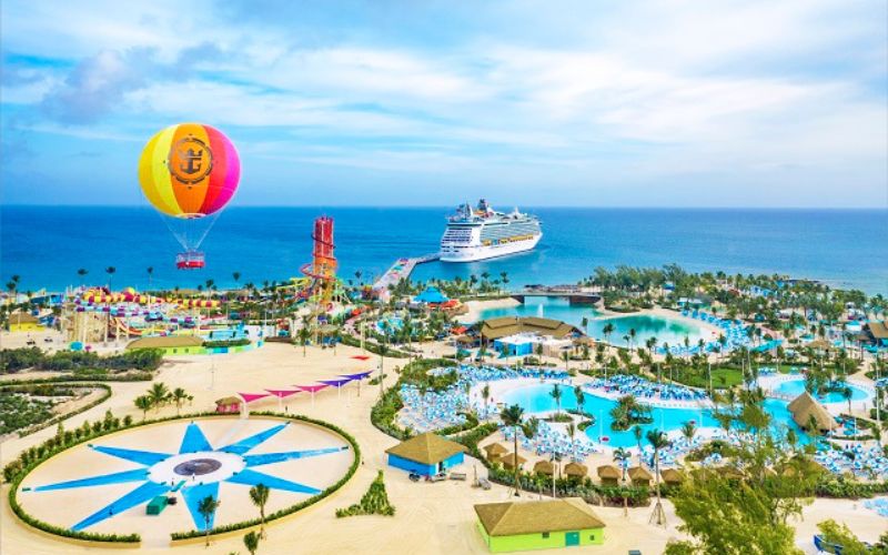 This image offers a vibrant aerial view of Perfect Day at CocoCay, a private island resort in the Bahamas operated by Royal Caribbean. The picture shows a colorful water park featuring a large swimming pool, water slides, and ample lounging areas. A hot air balloon offers rides above the island, providing panoramic views, while a cruise ship is moored nearby, emphasizing the island's role as a cruise destination. The entire scene is set against a backdrop of the turquoise Caribbean Sea and a blue sky, creating a lively and inviting vacation atmosphere.