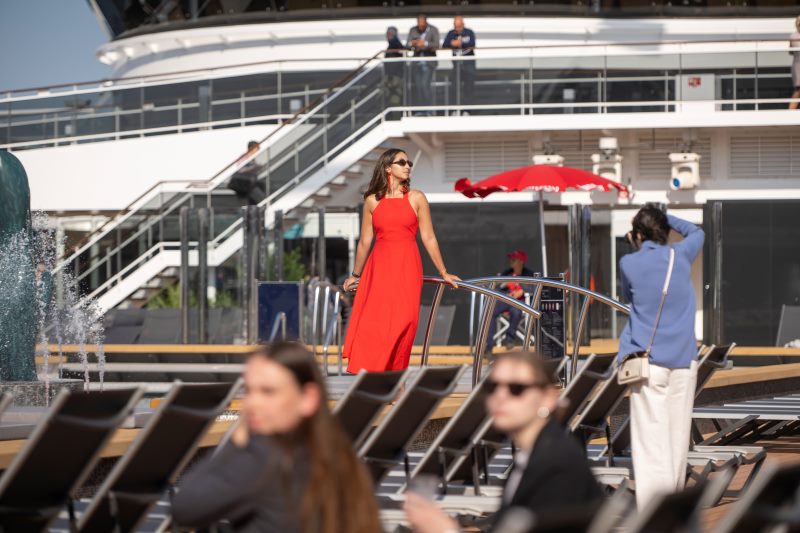 A photographer on a cruise ship captures a woman in a striking red dress posing on the deck. The woman stands gracefully by a railing, while other passengers relax on lounge chairs in the foreground. The ship's upper decks and architecture form the backdrop, and a few crew members are visible above, adding to the dynamic, lively scene.