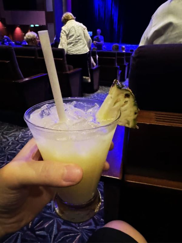 A hand holding a Piña Colada garnished with a pineapple slice inside a theater on the Royal Caribbean Ovation of the Seas. Passengers are seated, and a stage with blue lighting is visible in the background, creating a relaxed, pre-show atmosphere.