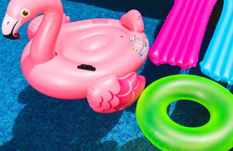 Colorful pool inflatables float on the water, including a pink flamingo, a green ring, and two long mats in pink and blue. The bright and playful items are set against the blue-tiled pool, suggesting a fun and relaxing time in the sun.