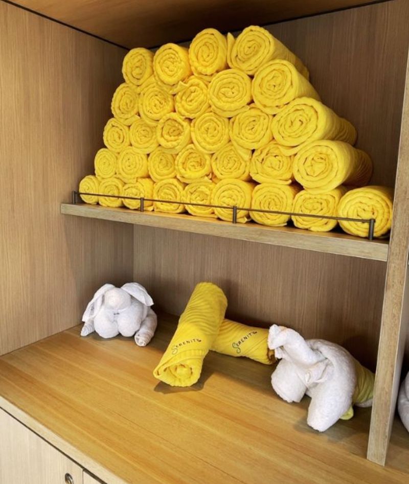Vibrant yellow pool towels neatly rolled and stacked on a shelf in a cruise ship spa, with whimsical white towel animals, including an elephant and rabbits, playfully arranged on the wooden cabinet below.
