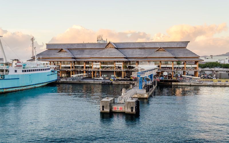 The bustling Port of Papeete in Tahiti, showcasing its unique blend of modern maritime architecture and tropical ambiance. The main building at the port, featured prominently in the photo, is constructed with a large, open-air design, typical of Pacific island architecture, which allows for natural ventilation and light. This structure is accompanied by wooden accents and a high, sloping roof, reflecting local styles. The calm sea in the foreground and the soft glow of the sunset contribute to a peaceful, picturesque setting. Various small boats and a larger ship docked nearby indicate the port's role as a key hub for both local and international maritime traffic, essential for both commerce and tourism in Tahiti.