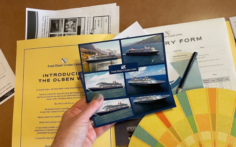 My hand holding a brochure from Fred. Olsen Cruise Lines with a collection of postcards featuring cruise ships, alongside a filled-out entry form and colorful brochures spread out on a table, indicating travel planning or booking for a cruise.