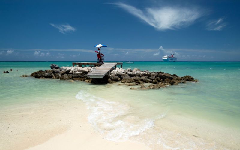 A lifeguard stands under a blue umbrella on a wooden pier at Princess Cays, vigilantly watching over swimmers in the shallow, crystal-clear turquoise waters. In the background, a cruise ship sits majestically on the horizon beneath a vast sky with wispy clouds.