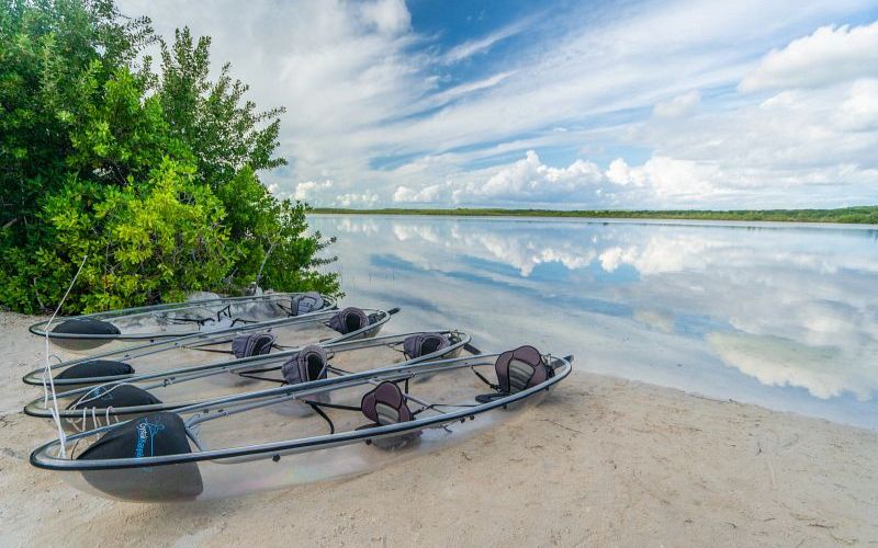 Transparent kayaks rest on the sandy edge of a calm lagoon at Princess Cays, ready for an adventure. The still waters mirror the sky and clouds above, creating a seamless blend of nature's elements, flanked by green foliage on one side.