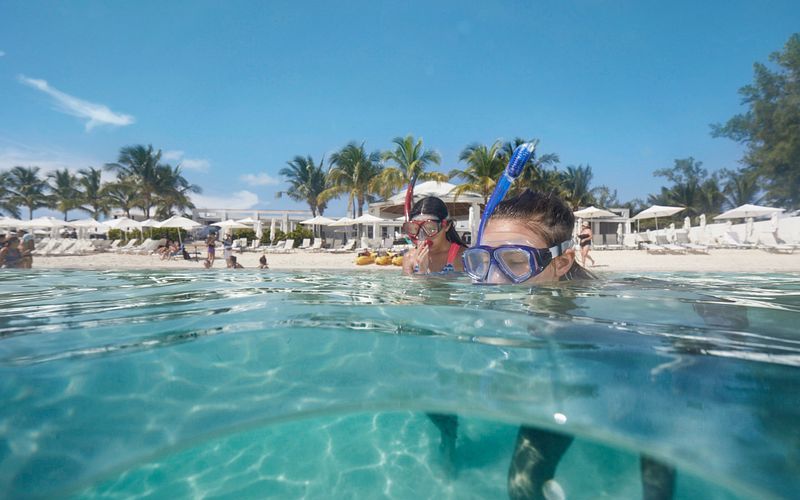 A close-up, split-level view of two kids snorkeling in the clear turquoise waters of Princess Cays, with one child's face partially submerged, looking at the camera through a snorkel mask. The beach resort in the background bustles with activity under a blue sky.