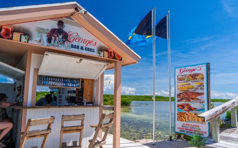 George's Bar & Grill, a quaint beachside hut at Princess Cays, welcomes guests with its laid-back ambiance. Patrons can enjoy local flavors under the shade, with a Bahamian flag waving proudly beside a menu board displaying an array of tempting dishes, all against a backdrop of serene lagoon waters.
