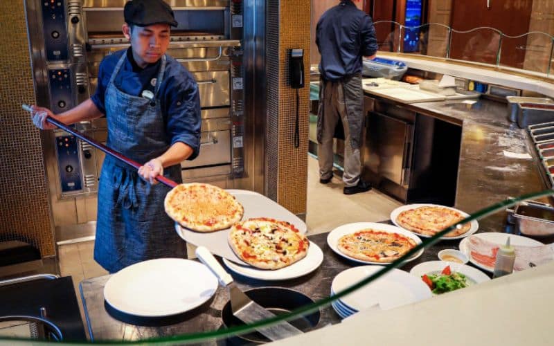 A chef on a Princess cruise ship prepares pizzas in a kitchen, placing a freshly baked pizza on a plate. Several other pizzas are ready to be served, and various ingredients and utensils are visible on the counter.