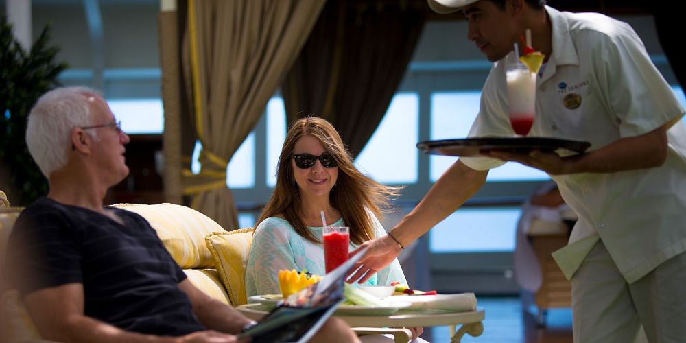This image depicts a relaxed scene aboard a cruise, featuring a man and woman lounging with drinks being served by a staff member. The woman smiles while enjoying a vibrant red cocktail, as the server brings another drink garnished with fruit. The cozy cabana-like setting adds an air of luxury and comfort, highlighting the cruise's personalized service.