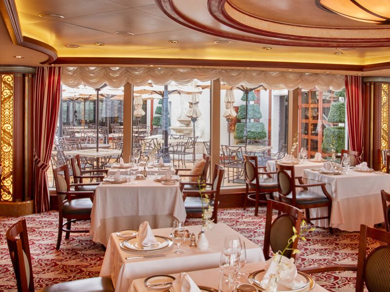 Elegant dining setting inside the Princess Grill Restaurant on board Cunard's Queen Elizabeth cruise ship. The photo depicts a well-appointed area with tables dressed in white linen, fine china, and crystal glassware. Red patterned carpet, ornate golden lighting, and large windows offering a view to the outdoor seating area create an ambiance of classic luxury.