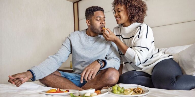 This image features a couple enjoying room service aboard a cruise, sitting on a neatly made bed. The woman playfully feeds the man while they share a spread of fresh fruit, vegetables, and other light snacks on white plates. The cozy, relaxed setting highlights the intimate and personalized dining experience offered in their cabin.