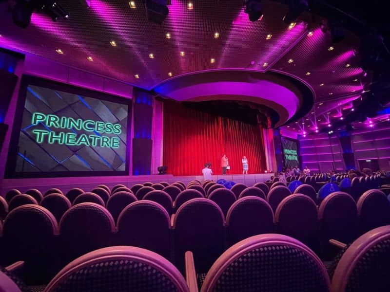The Princess Theatre on a Princess cruise ship features a large stage with a red curtain and two screens displaying the theater's name. The seating area is filled with rows of cushioned chairs under a ceiling illuminated with pink and purple lighting, creating a vibrant atmosphere.