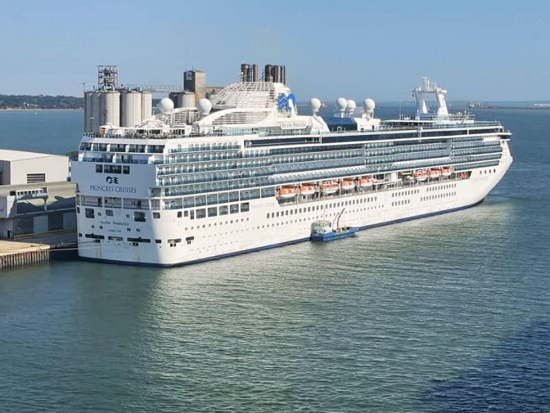 A large Princess Cruises ship, the Island Princess, is docked at a port. The ship is white with multiple decks and lifeboats visible along the side, with industrial buildings and the ocean in the background under a clear blue sky.