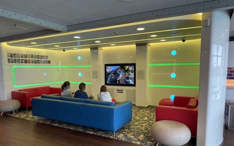 Kids club room on Princess Cruise Line, featuring colorful sofas and a large TV where children are playing video games. The room has modern design elements with illuminated green accents on the walls, creating a fun and engaging environment for young passengers.