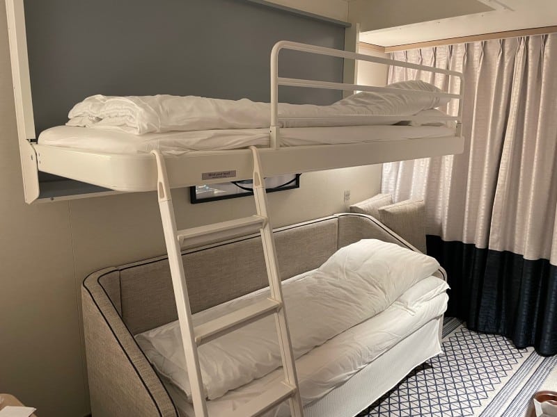 This image shows a neatly arranged small room with a bunk bed configuration, featuring a lower couch that likely converts into a bed and an upper bunk accessible by a ladder. The room has a neutral color palette, with gray walls and white bedding, and is complemented by soft curtain drapes that suggest a cozy and compact living space, possibly within a cruise ship or a budget hotel room designed to efficiently accommodate multiple guests.