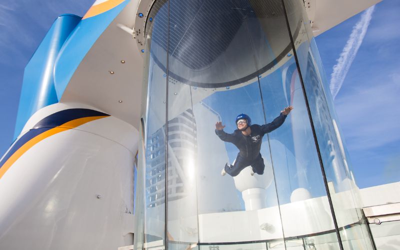 A person in a blue flight suit and helmet joyfully experiences simulated skydiving in the RipCord by iFLY, a vertical wind tunnel aboard the Quantum of the Seas cruise ship, set against a backdrop of clear blue skies.