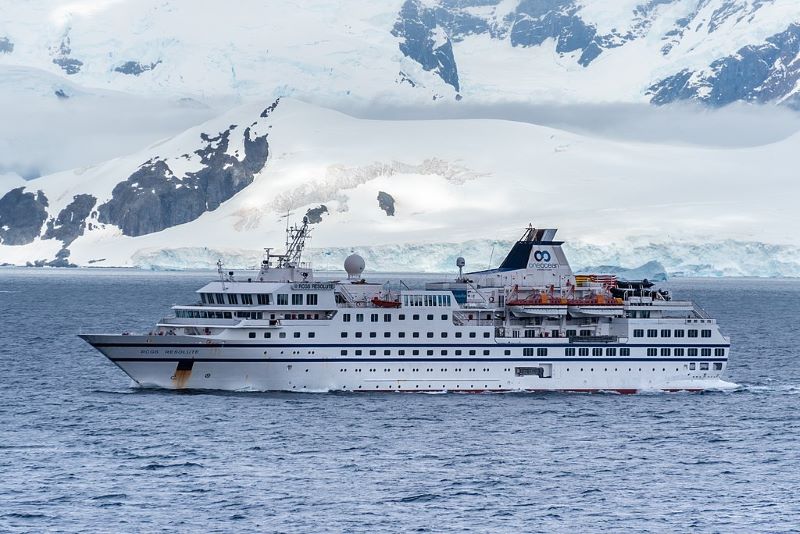 RCGS Resolute expedition cruise ship navigating through icy waters with a backdrop of dramatic Antarctic scenery. The ship, painted white with a distinctive blue and orange trim on the hull, contrasts strikingly against the rugged, snow-covered mountains and icy shores in the background. Equipped with advanced navigational and satellite technology on its upper decks, the vessel is clearly designed for voyages in challenging polar environments.