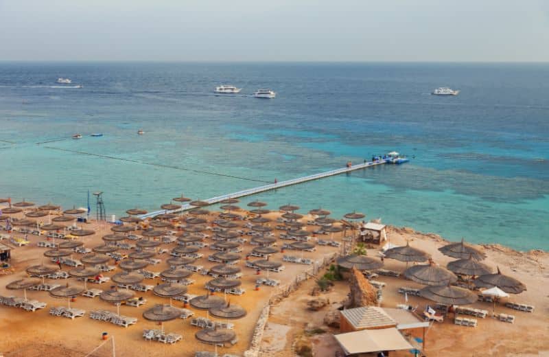 This image captures a bustling beach scene along the Red Sea coast in Sharm el-Sheikh, Egypt. The beach is densely lined with rows of parasols and sun loungers, offering a popular spot for relaxation and sunbathing. A long pier extends into the clear turquoise waters, where several boats are moored, suggesting opportunities for water activities like snorkeling or diving. The backdrop features a rocky desert landscape, emphasizing the unique geographical mix of sea and arid terrain typical of this renowned tourist destination.