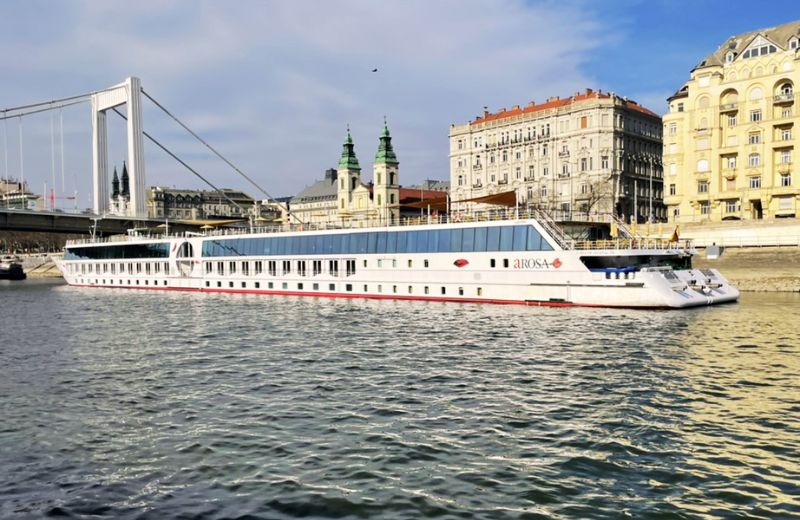 A serene view of an A-Rosa river cruise ship docked along the waterfront in a picturesque European city. The scene is set against an urban backdrop with elegant, historic buildings and a distinctive white suspension bridge, suggesting the location could be along a well-known river in Europe, possibly the Danube in Budapest. The cruise ship, long and white with red accents, is designed for navigating the narrower and shallower waters of riverways, offering passengers a close-up view of cityscapes and landscapes. The calm water and clear skies contribute to a peaceful and inviting travel atmosphere, ideal for exploring the cultural and architectural beauty along European rivers.