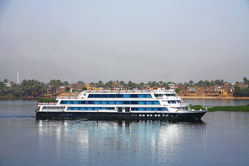The Riviera Travel river cruise ship is captured mid-voyage on the Nile, its blue and white exterior crisp against the backdrop of the Egyptian landscape. Passengers on the upper deck appear to be enjoying the panoramic views, with the rural shoreline and palm trees providing a tranquil setting for their journey.