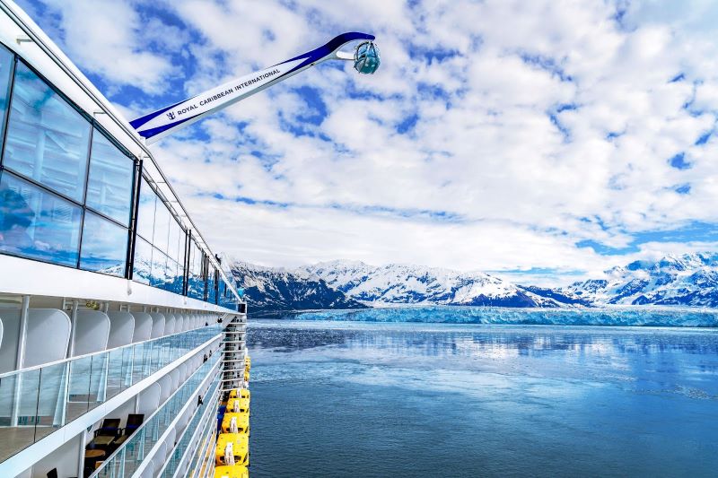 A Royal Caribbean cruise ship in Alaska with the North Star observation capsule extended, offering panoramic views over the icy Alaskan waters and snow-capped mountains under a vast blue sky with scattered clouds.