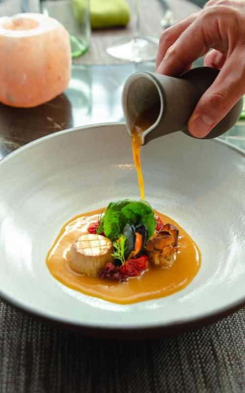 An elegant dish being meticulously plated, with a hand pouring a vibrant orange sauce over beautifully arranged seafood and garnishes, in a chic dining setting on a cruise ship, exemplifying fine dining at sea.
