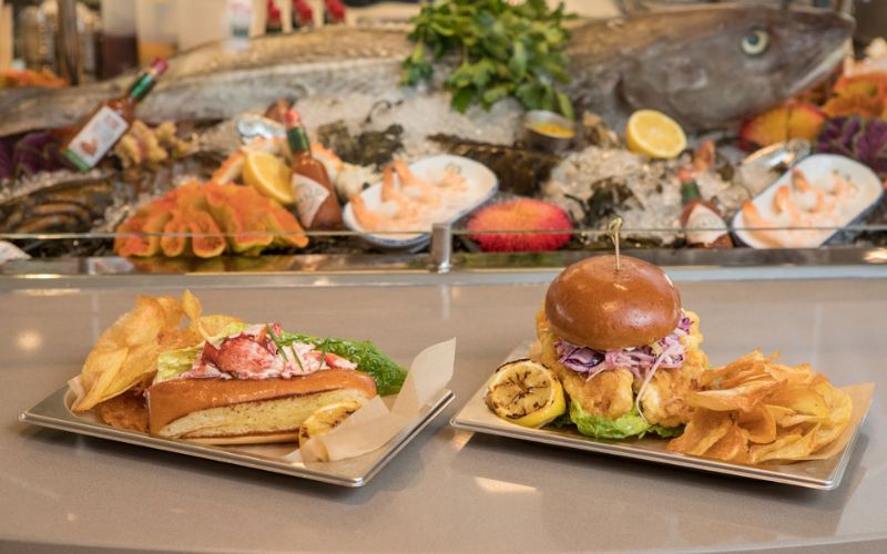 A close-up of two gourmet dishes served on a cruise: a lobster roll with a side of chips on the left, and a crispy chicken burger with plantain chips on the right. In the background, a seafood display with a whole fish, shrimp, and lemons creates an inviting atmosphere for dining.