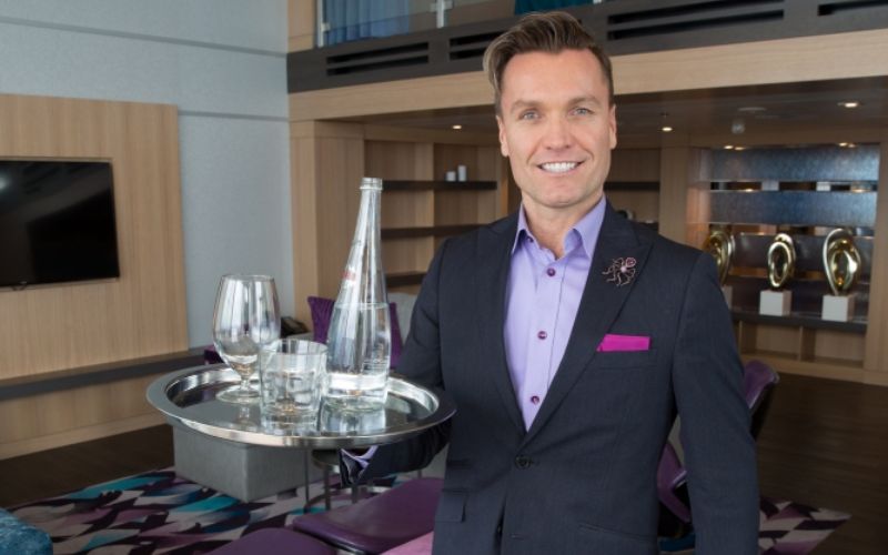 A cheerful Royal Genie, Royal Caribbean's personalized concierge service, stands ready in a luxury suite, holding a tray with sparkling water and glasses, offering an elevated, tailor-made guest experience onboard the cruise ship.