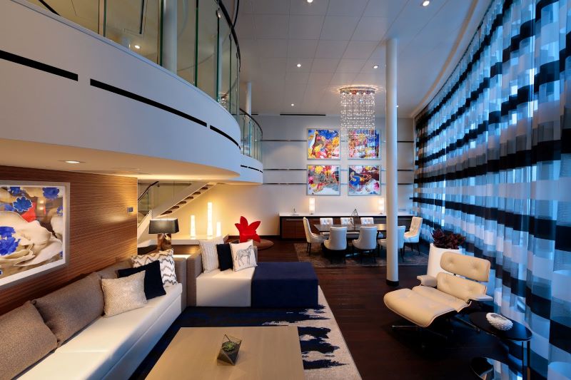 Interior view of the Royal Loft Suite on Royal Caribbean's Anthem of the Seas, displaying a two-level luxury space with sleek, modern furnishings, a dynamic blue light wall, and vibrant pop art, all under the glow of a chic chandelier.